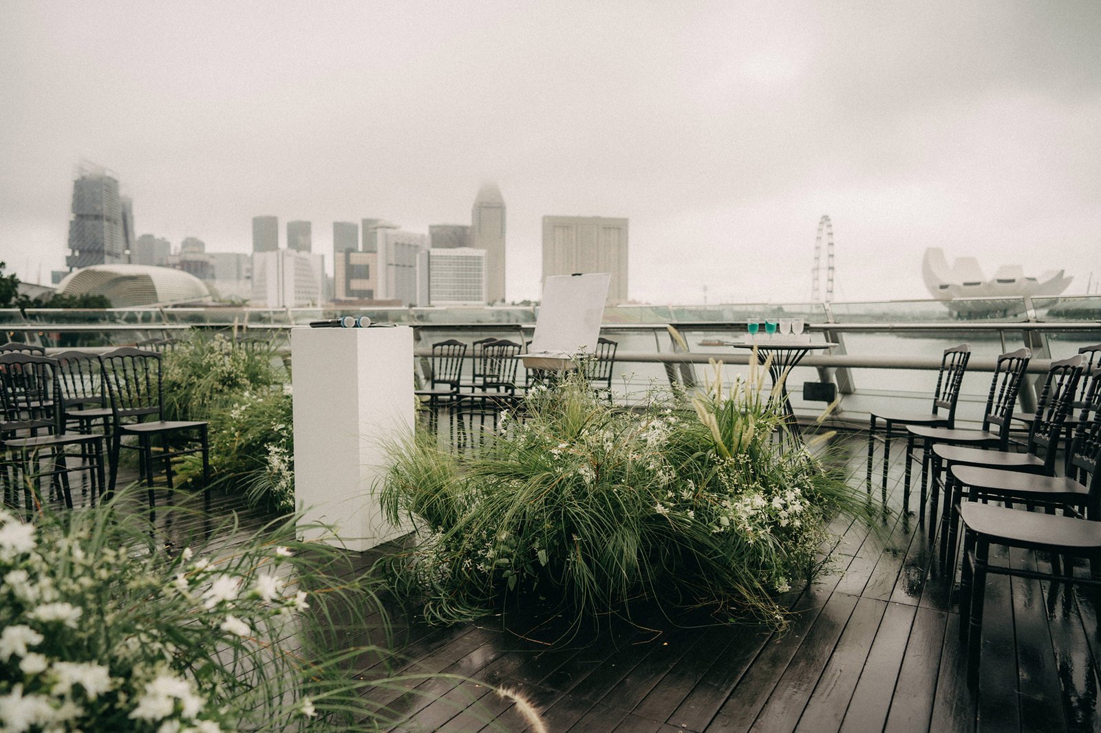 Outdoor wedding ceremony setup at Monti at 1-Pavilion, Singapore, with chairs arranged on a wet wooden deck overlooking a misty city skyline. Lush greenery and white flowers adorn the aisle, creating a romantic atmosphere for a wedding photography session.