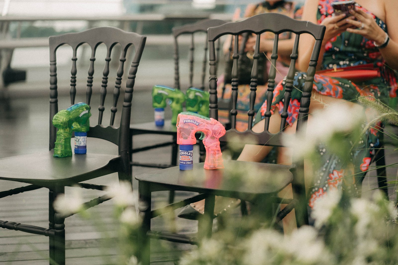 Pink and green dinosaur-shaped bubble guns sit on chairs at a wedding ceremony at Monti, Singapore, with guests in colorful attire blurred in the background.