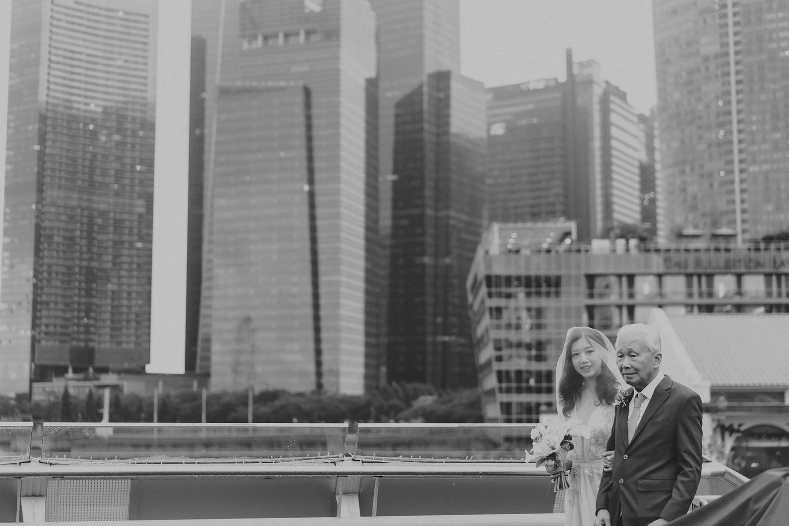 Black and white wedding photography at Monti at 1-Pavilion, Singapore: A smiling bride in a veil and a groom in a suit walk together with a city skyline in the background.