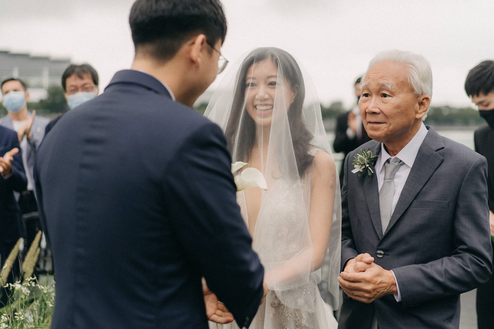 A joyful bride in a veil smiles at her groom during a wedding ceremony at Monti at 1-Pavilion, Singapore, with her father looking on.