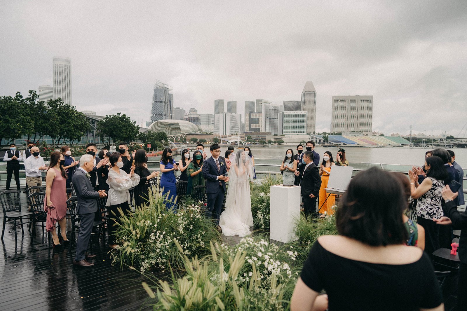 Monti wedding in Singapore: Bride and groom exchange vows outdoors with guests watching, with city skyline and marina in the background.