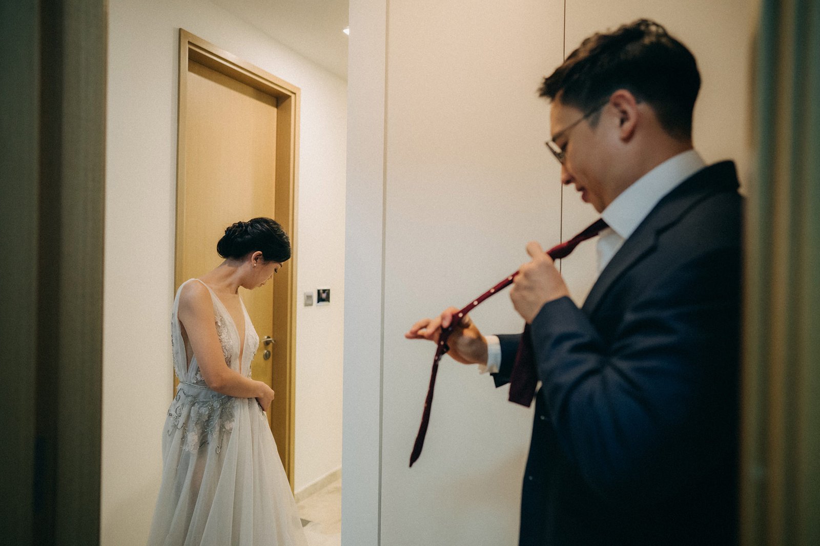 Bride in a flowing gown looks down as groom in a suit ties his maroon tie, preparing for their Monti at 1-Pavilion wedding in Singapore, captured by wedding photography.