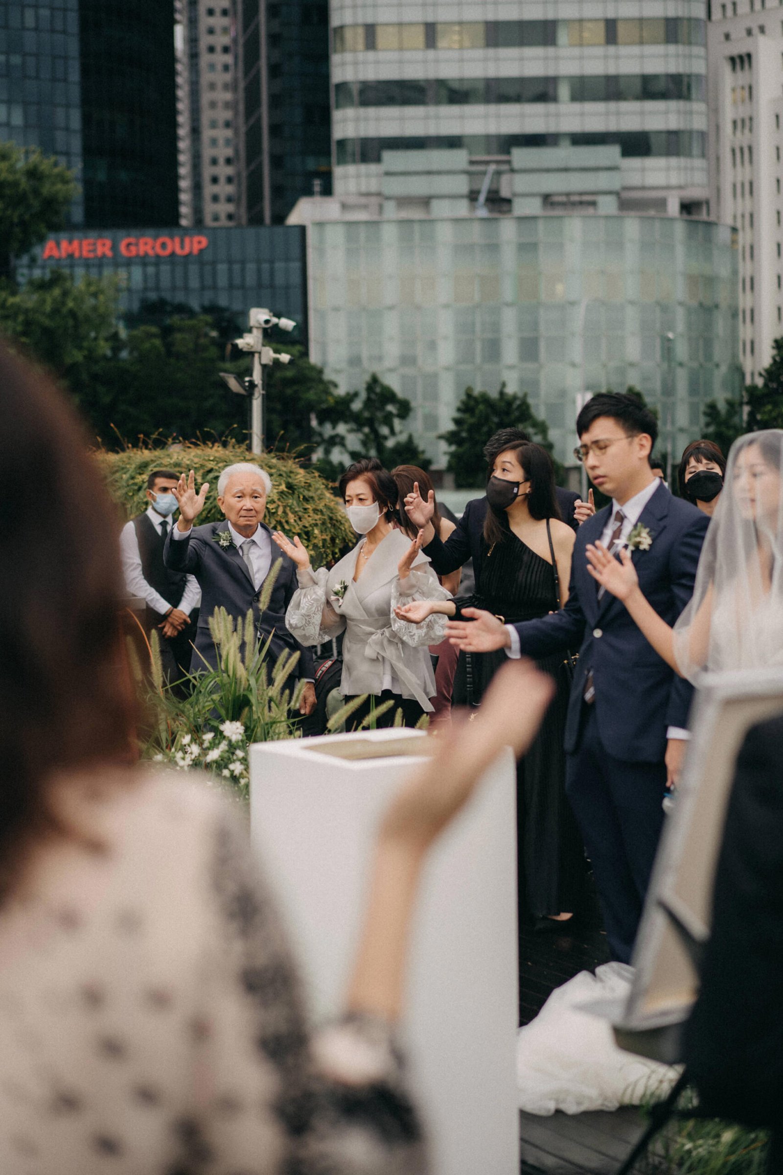 Wedding ceremony at Monti at 1-Pavilion, Singapore. An elderly man in a suit gestures during an outdoor event, with guests in formal attire and masks in the background. Modern buildings form the backdrop.