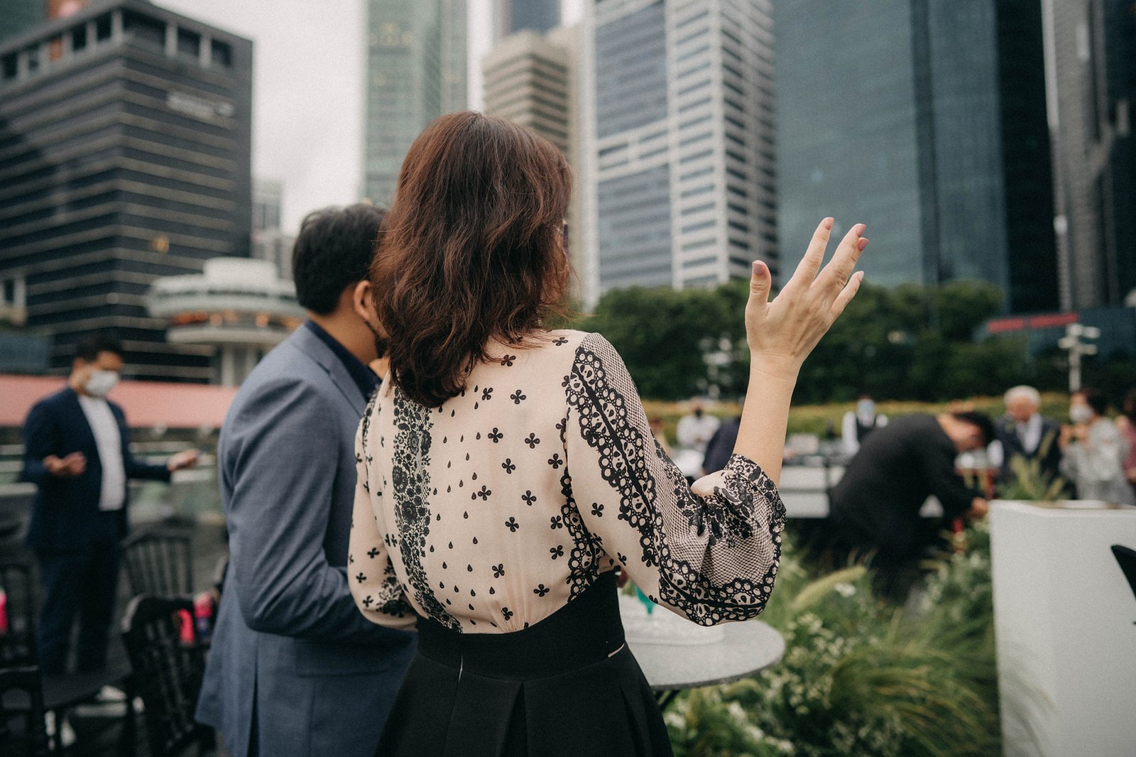 A woman in a patterned blouse and black skirt gestures with her hand, facing away from the camera at an outdoor event at Monti at 1-Pavilion, Singapore. A man in a blue suit stands beside her, with the city skyline in the background.