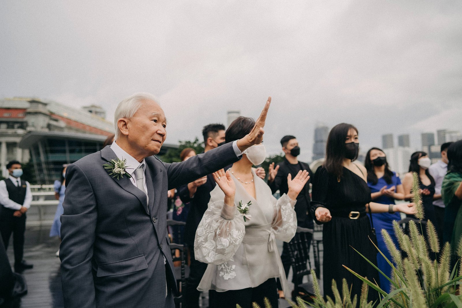 Elderly man in a grey suit with a boutonniere raises his hand at a wedding ceremony at Monti, Singapore, with guests in formal attire and masks in the background. Monti at 1-Pavilion wedding photography.