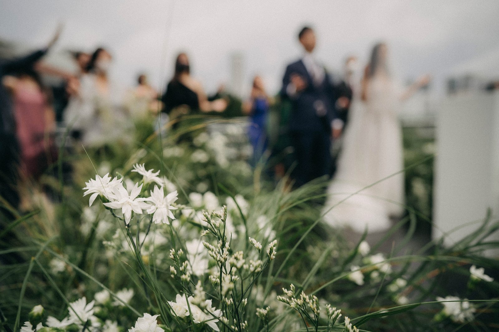 A wedding ceremony at Monti at 1-Pavilion Singapore, with a blurred foreground of white flowers and greenery, and the bride and groom walking down the aisle in the background.