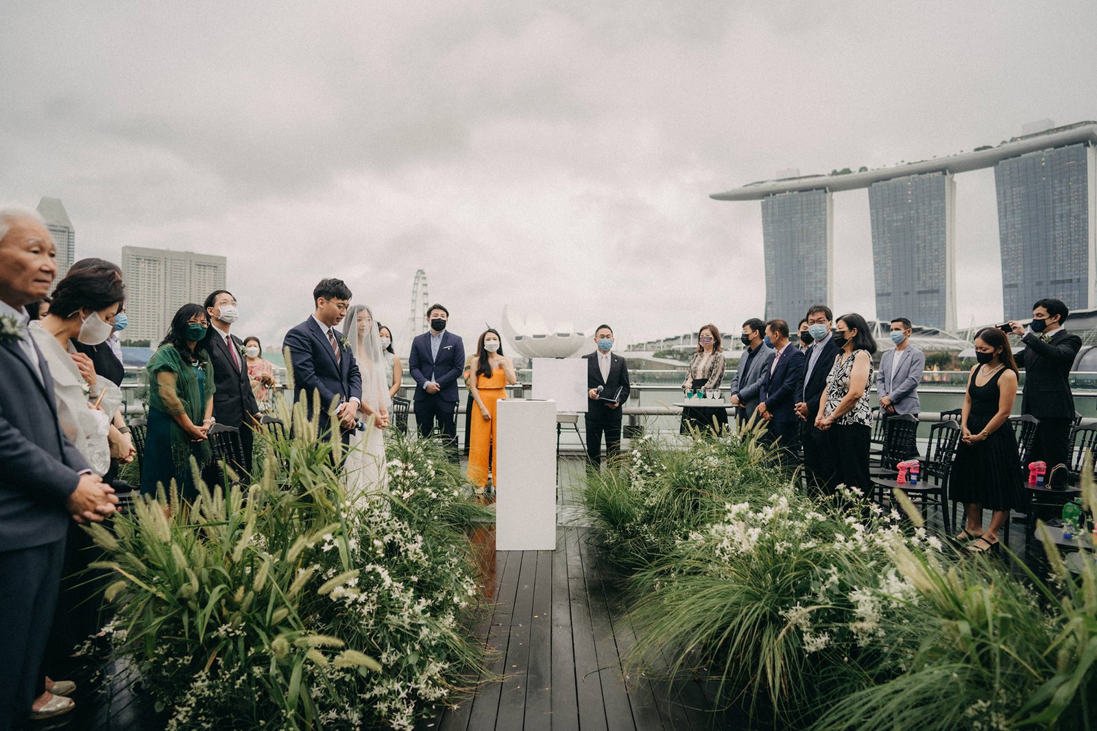 A modern wedding ceremony at Monti at 1-Pavilion, Singapore, with a bride and groom at a white altar, surrounded by guests and lush greenery, against a backdrop of the city skyline.