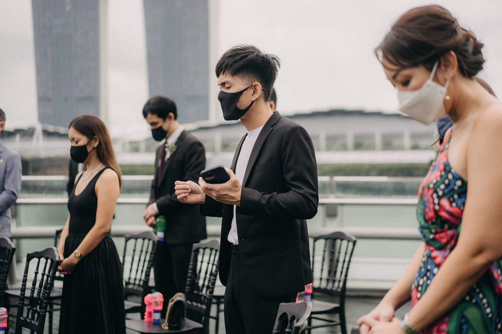 Guests in masks stand during an intimate wedding ceremony at Monti at 1-Pavilion, Singapore. The bride's guests wear elegant black, while the groom's side is in formal attire, all facing forward with solemn expressions.