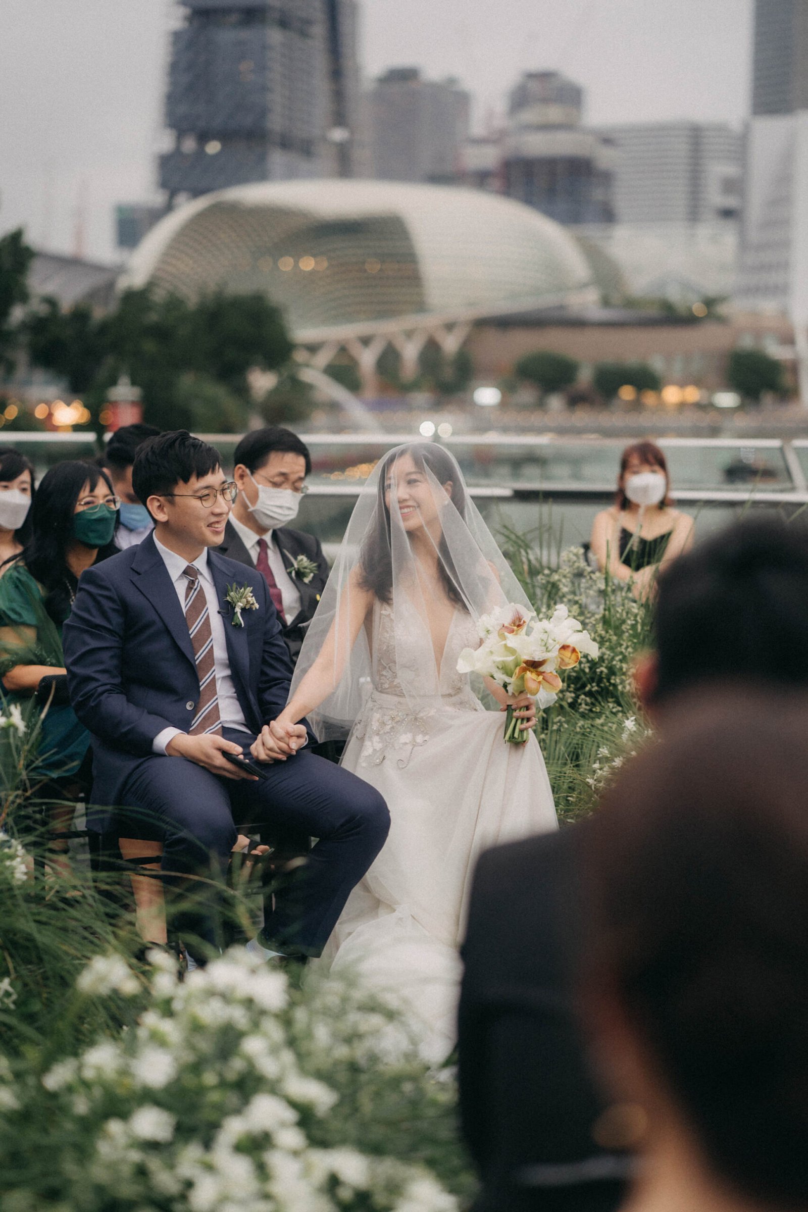 A bride and groom hold hands during their wedding ceremony at Monti at 1-Pavilion, Singapore, with the city skyline in the background. The bride wears a flowing white gown and veil, while the groom is in a navy suit.