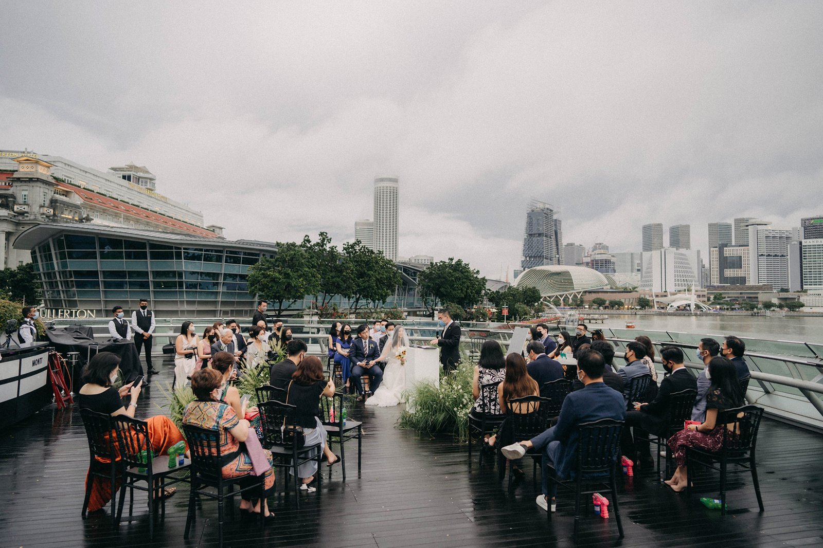 Monti at 1-Pavilion wedding in Singapore: A bride and groom exchange vows on an outdoor deck overlooking the city skyline, with guests seated on either side.