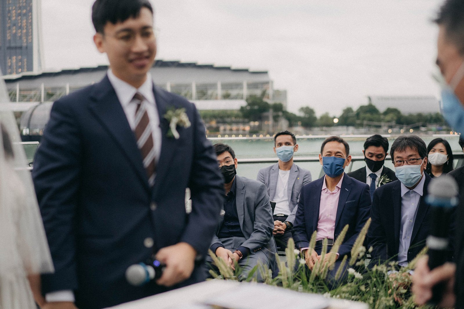 Groom in a dark suit and mask smiles during a wedding ceremony at Monti at 1-Pavilion, Singapore, with guests seated in the background.