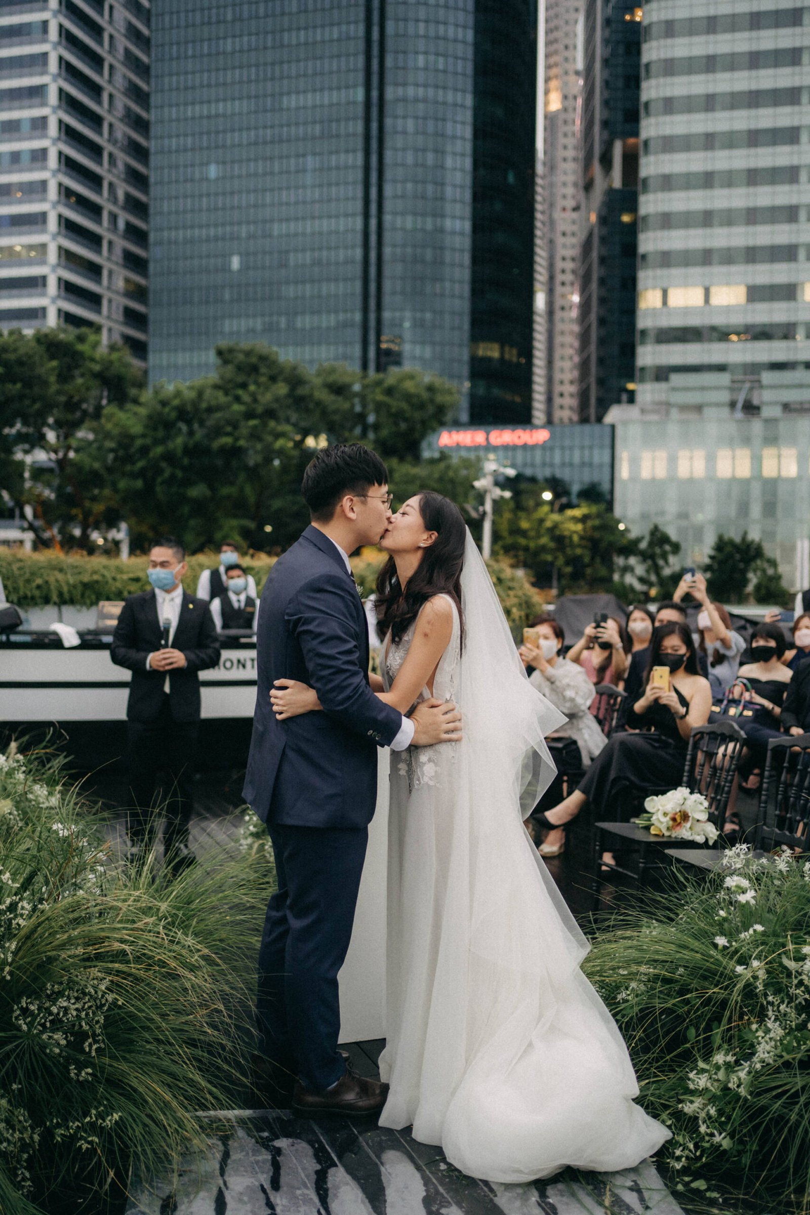 Wedding ceremony at Monti at 1-Pavilion, Singapore: Bride and groom kiss at their outdoor wedding, with city skyscrapers and guests in the background.