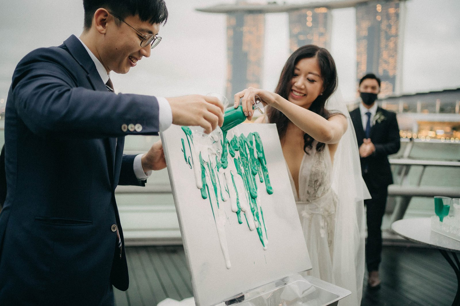 A joyful bride and groom pour green paint onto a white canvas during a creative wedding ceremony at Monti at 1-Pavilion, Singapore, with the city skyline in the background.
