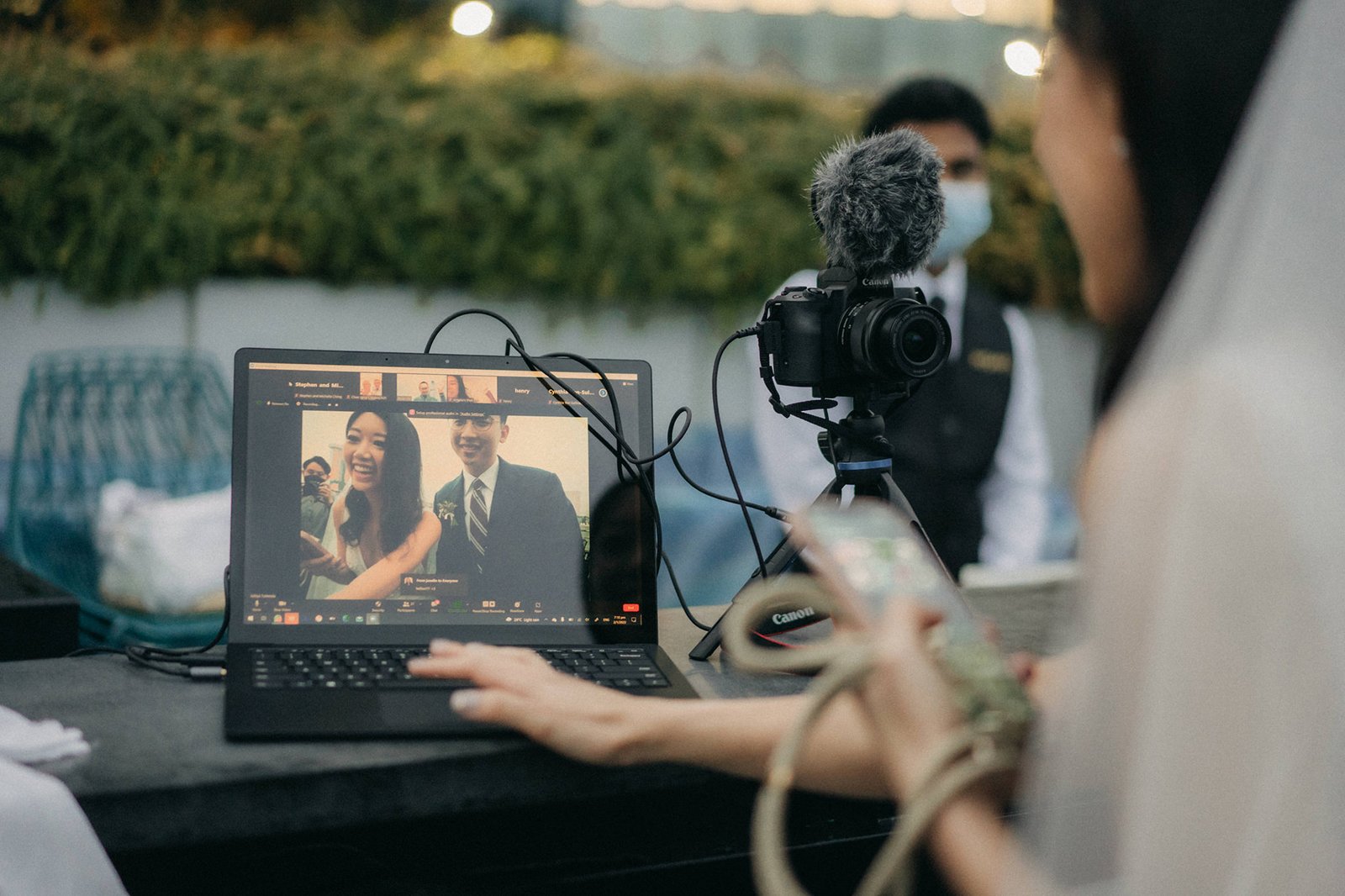 A bride in a veil smiles while looking at a laptop screen displaying a video call of a wedding couple, set against the backdrop of a Monti at 1-Pavilion wedding in Singapore. A camera on a tripod is also visible.