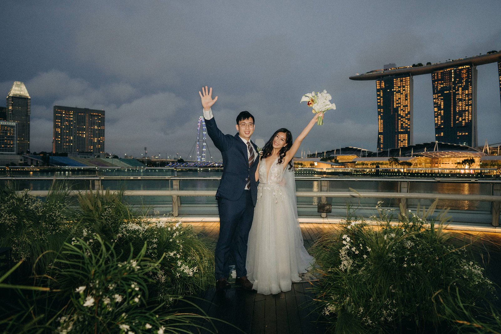 Wedding couple, LK and Sophie, wave happily on their wedding day at Monti at 1-Pavilion in Singapore, with the city skyline at dusk in the background.