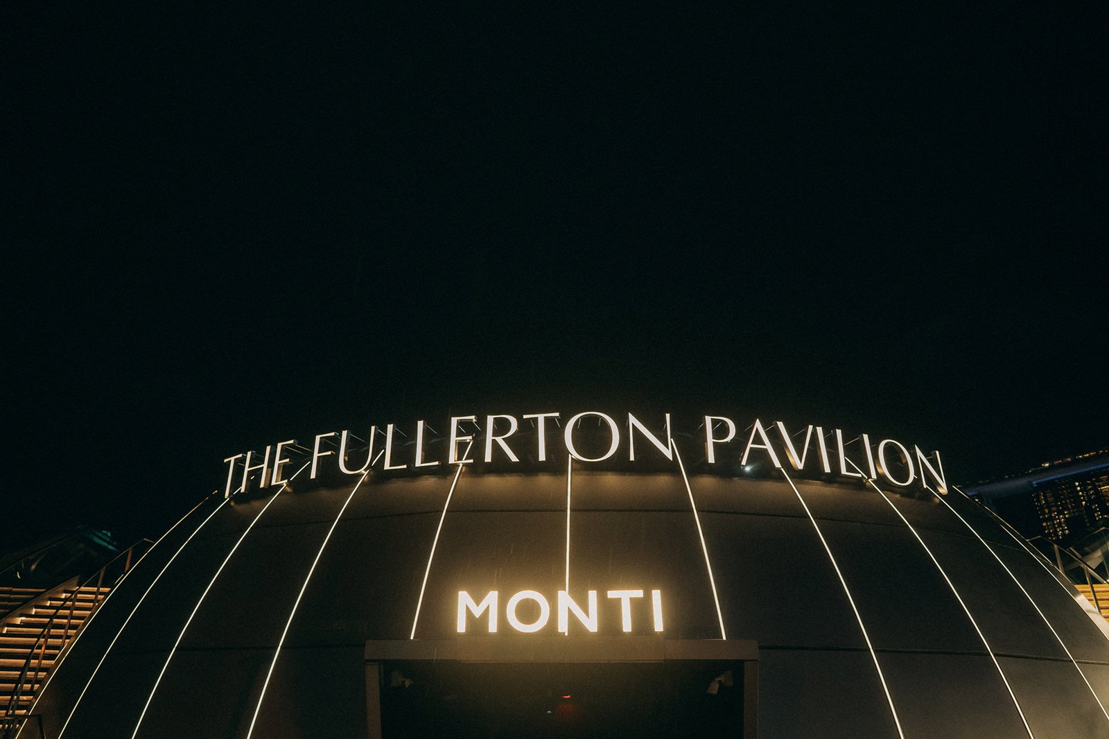 The Fullerton Pavilion at night, with bright neon signs reading "THE FULLERTON PAVILION" and "MONTI" illuminated against a dark sky, hinting at a wedding photography venue in Singapore.