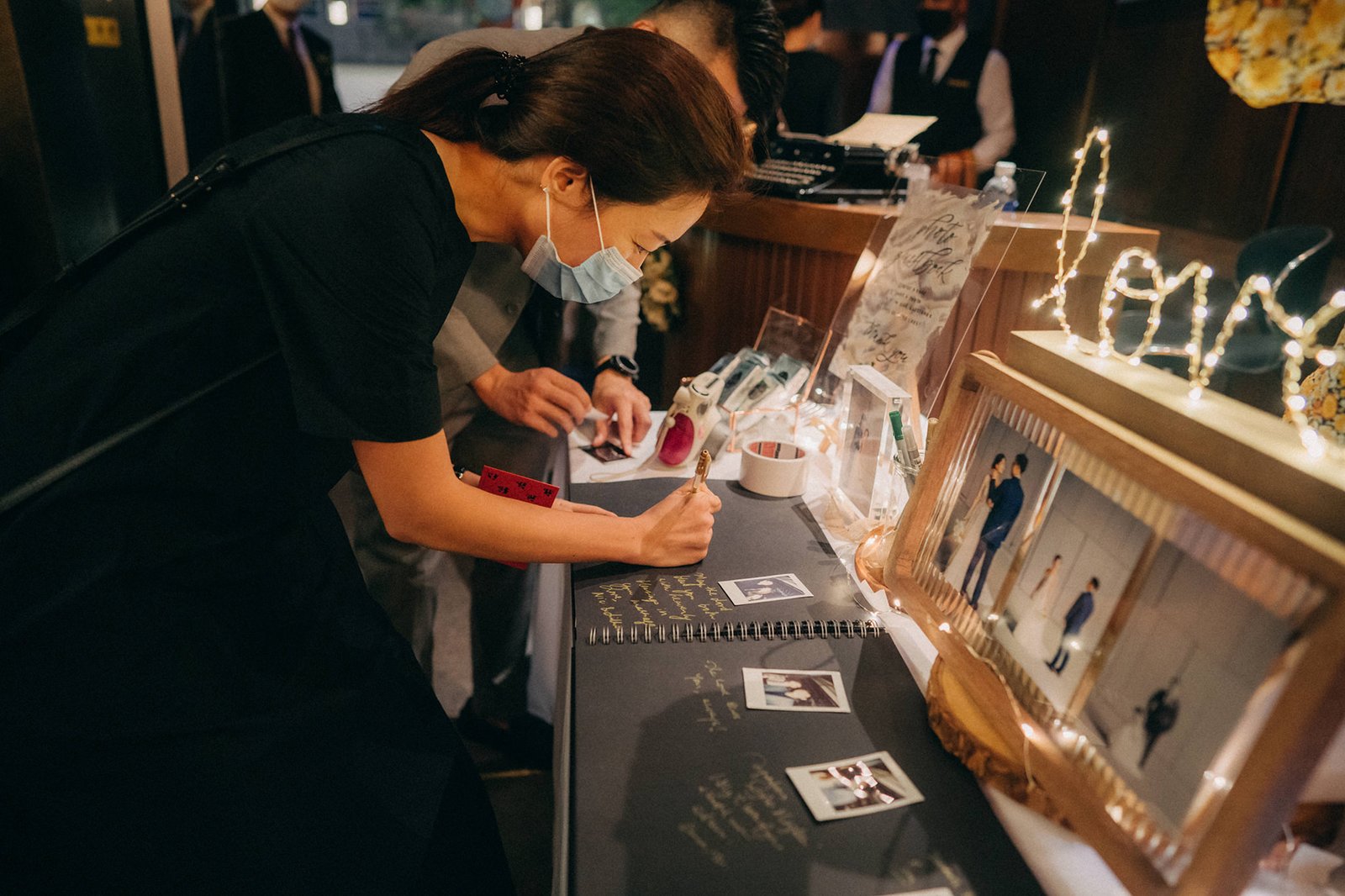 A woman in a black dress signs a guestbook at a wedding reception at Monti at 1-Pavilion, Singapore, with photos and fairy lights in the background.