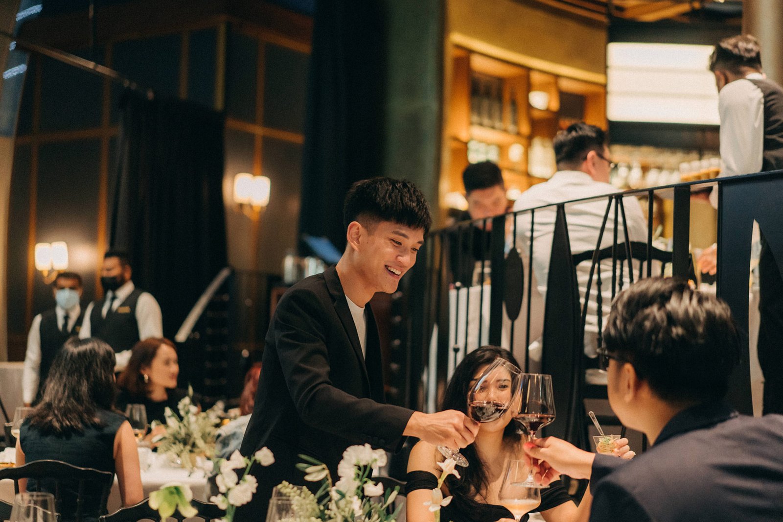 A smiling groom in a black suit toasts with a glass of red wine at a wedding reception at Monti at 1-Pavilion, Singapore, with guests and servers in the background.