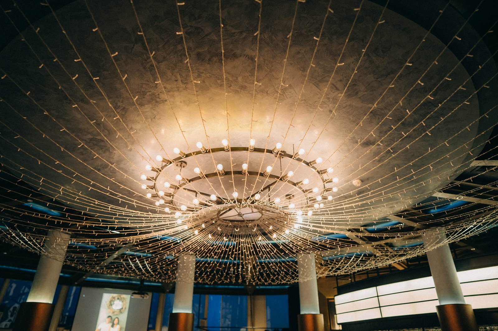 A stunning circular chandelier adorned with fairy lights hangs from a textured ceiling at Monti, a wedding venue in Singapore. Fairy lights cascade downwards, creating a magical atmosphere.