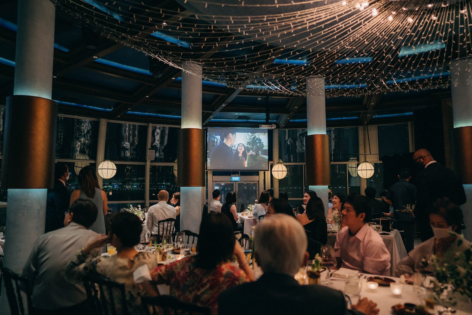 Wedding reception at Monti at 1-Pavilion, Singapore, with guests seated at round tables under a net-like ceiling adorned with lights. A large screen displays a couple.