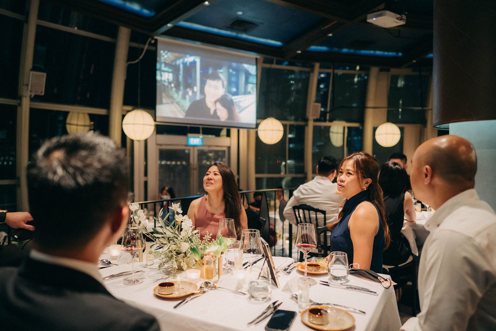 Guests seated at a wedding reception dinner at Monti at 1-Pavilion, Singapore, with a projection screen in the background.