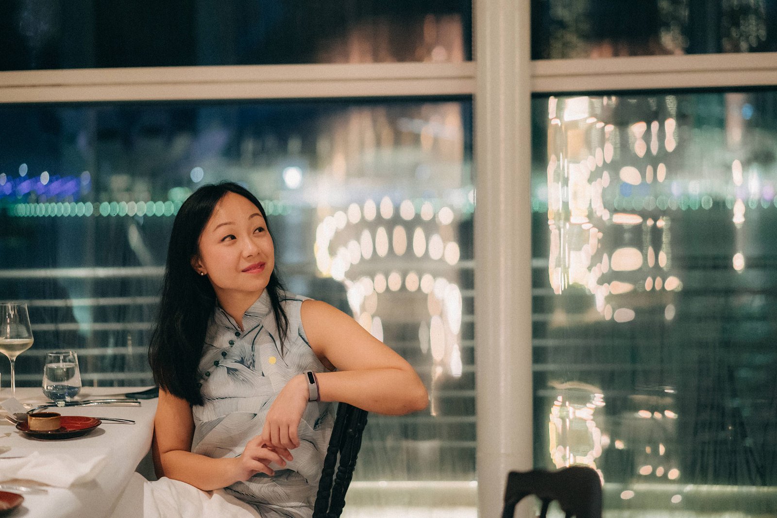 A woman with dark hair smiles and looks away from the camera while seated at a table with a view of city lights at Monti, Singapore, a venue for wedding photography.