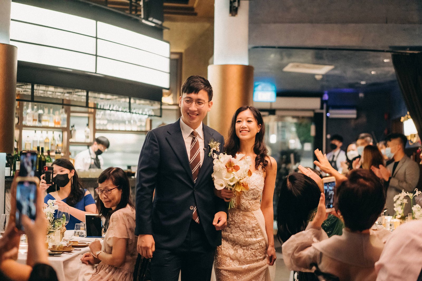 Newlyweds LK and Sophie smile as they walk through a wedding reception at Monti at 1-Pavilion, Singapore, with guests in the background capturing the moment. Monti at 1-Pavilion wedding Singapore photography.