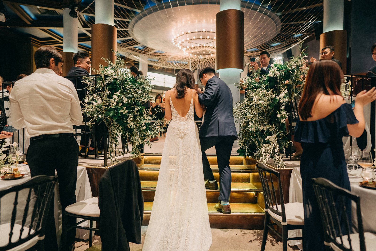 Bride and groom ascend stairs at a Monti at 1-Pavilion wedding in Singapore, surrounded by guests and lush greenery. Photography captures a romantic moment.