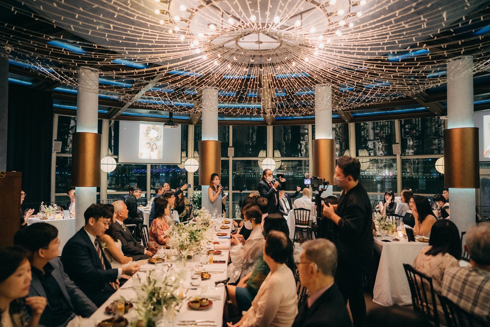 A wedding reception at Monti at 1-Pavilion, Singapore, with guests seated at tables under a dazzling ceiling of fairy lights. A woman is speaking at a microphone, with a projection screen displaying a couple in the background.