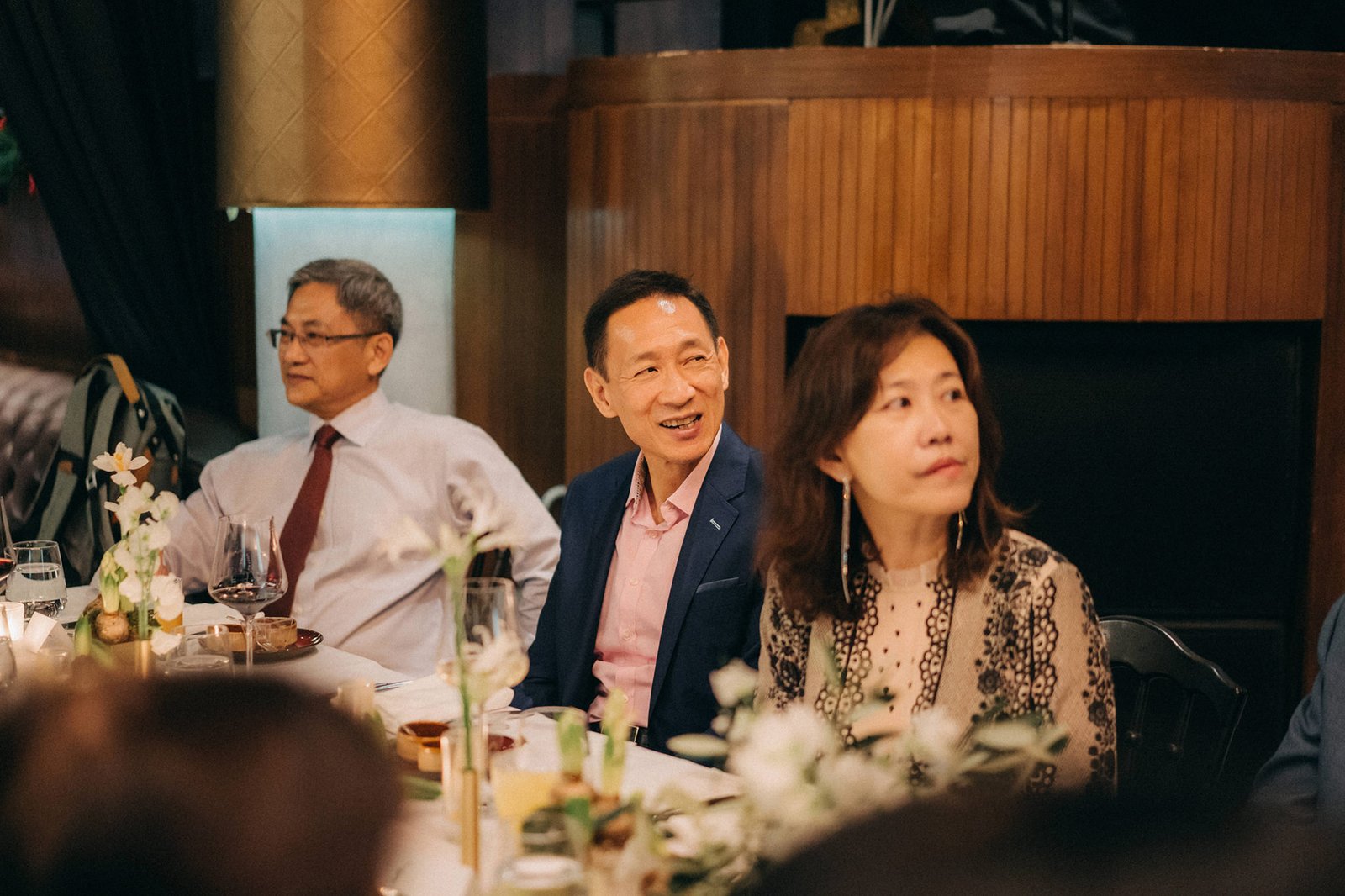 Guests at a Monti at 1-Pavilion wedding in Singapore, including a man in a navy suit and pink shirt, and a woman in a patterned top, smile and look on during a reception event, captured by wedding photography.