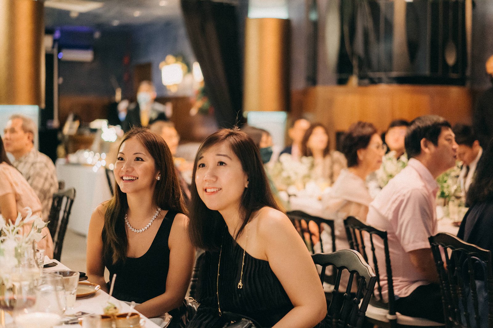 Two smiling women in elegant black dresses attend a wedding reception at Monti at 1-Pavilion, Singapore, with guests in the background.