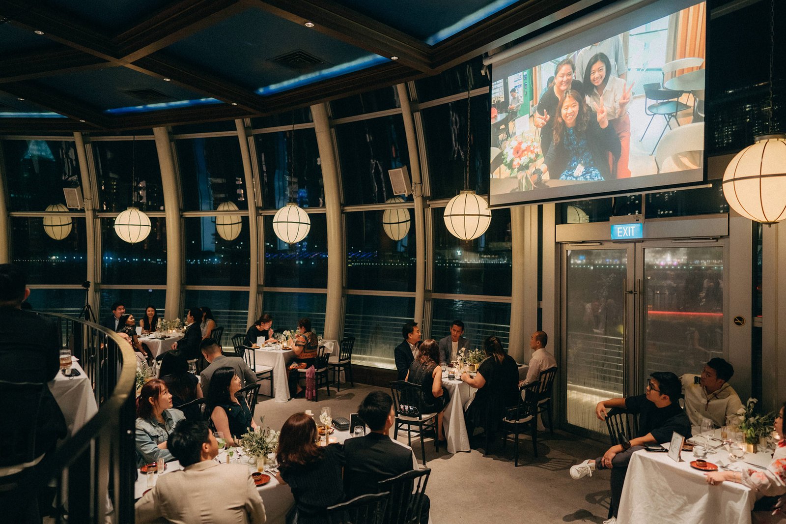 A wedding reception at Monti at 1-Pavilion, Singapore, with guests seated at tables under a projector screen showing a photo of the wedding couple. The elegant venue features large windows overlooking the city at night.