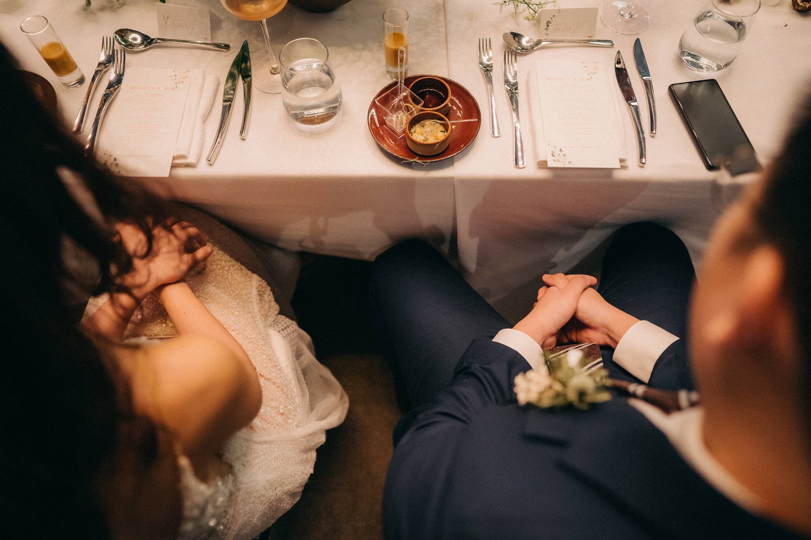 A bride and groom hold hands across a wedding reception table at Monti at 1-Pavilion, Singapore, with place settings and menus visible. Monti at 1-Pavilion wedding photography.