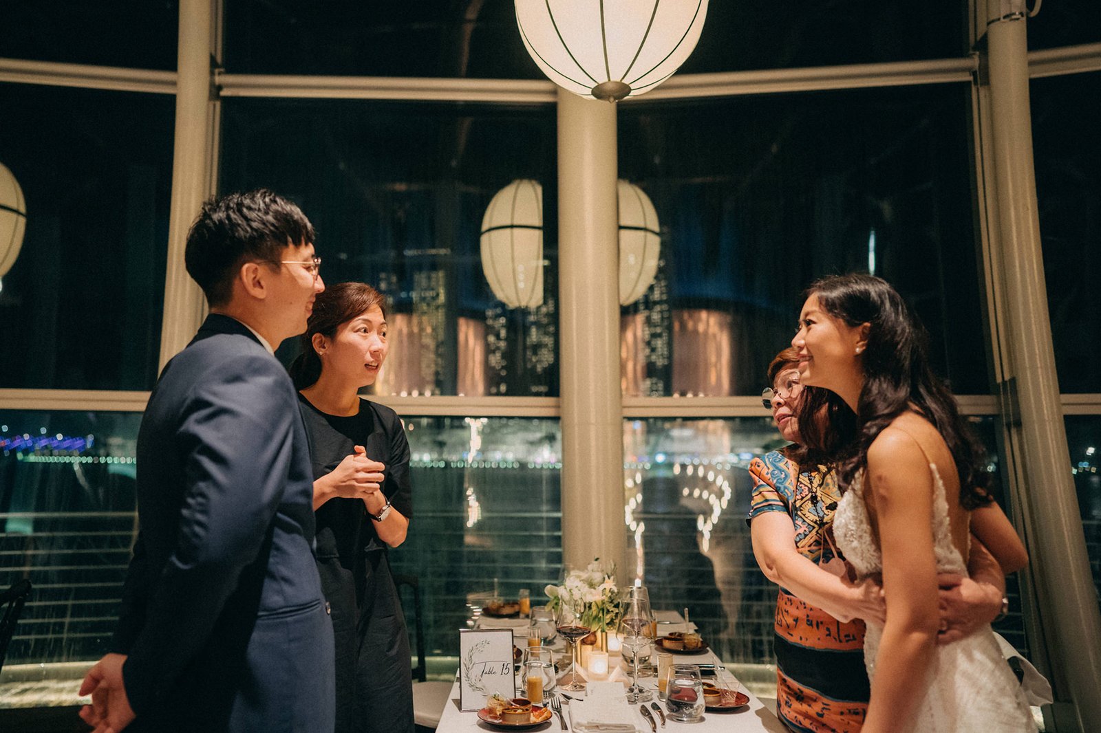 A wedding photography moment at Monti at 1-Pavilion, Singapore, where a bride in a white gown embraces a woman, while a groom in a suit and another woman converse near a table set for a reception.