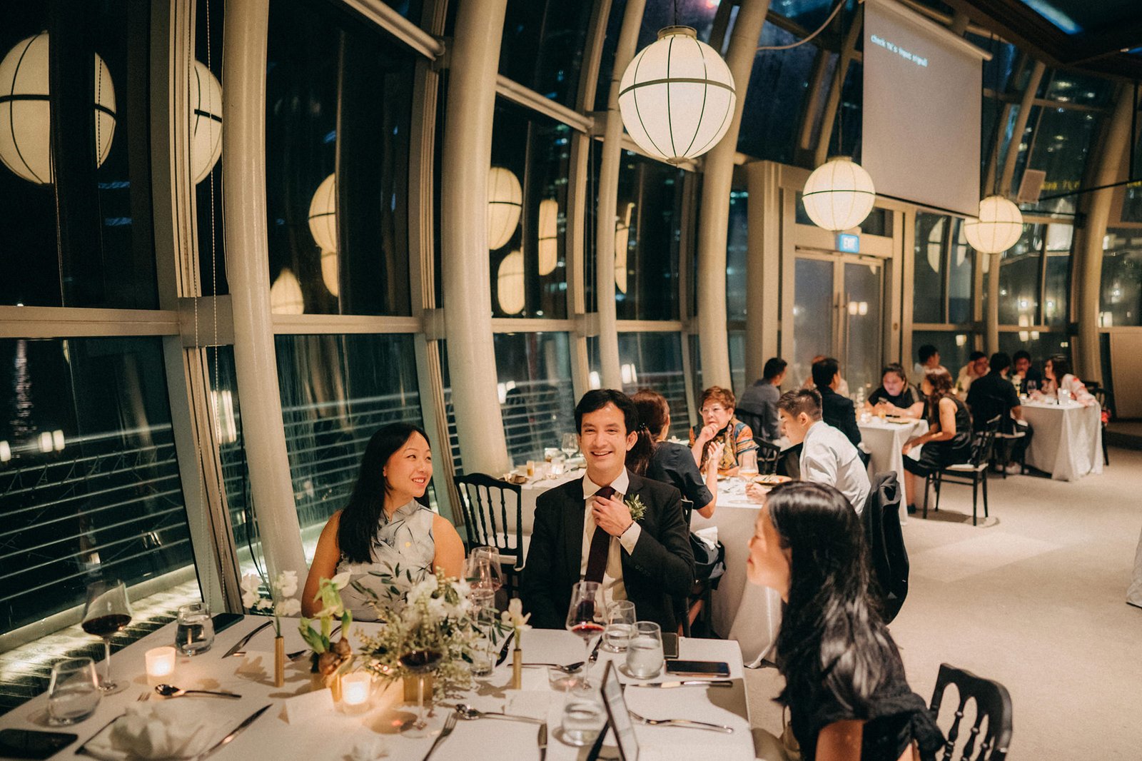 A wedding reception at Monti at 1-Pavilion, Singapore, with guests seated at tables. A couple smiles at each other in the foreground, with city lights visible through the large windows in the background.