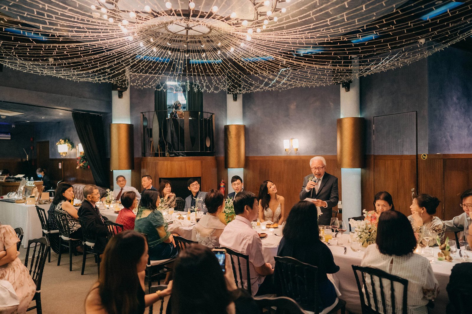 A wedding reception at Monti at 1-Pavilion, Singapore, with guests seated at round tables under a canopy of fairy lights. A man is giving a speech at the head table.