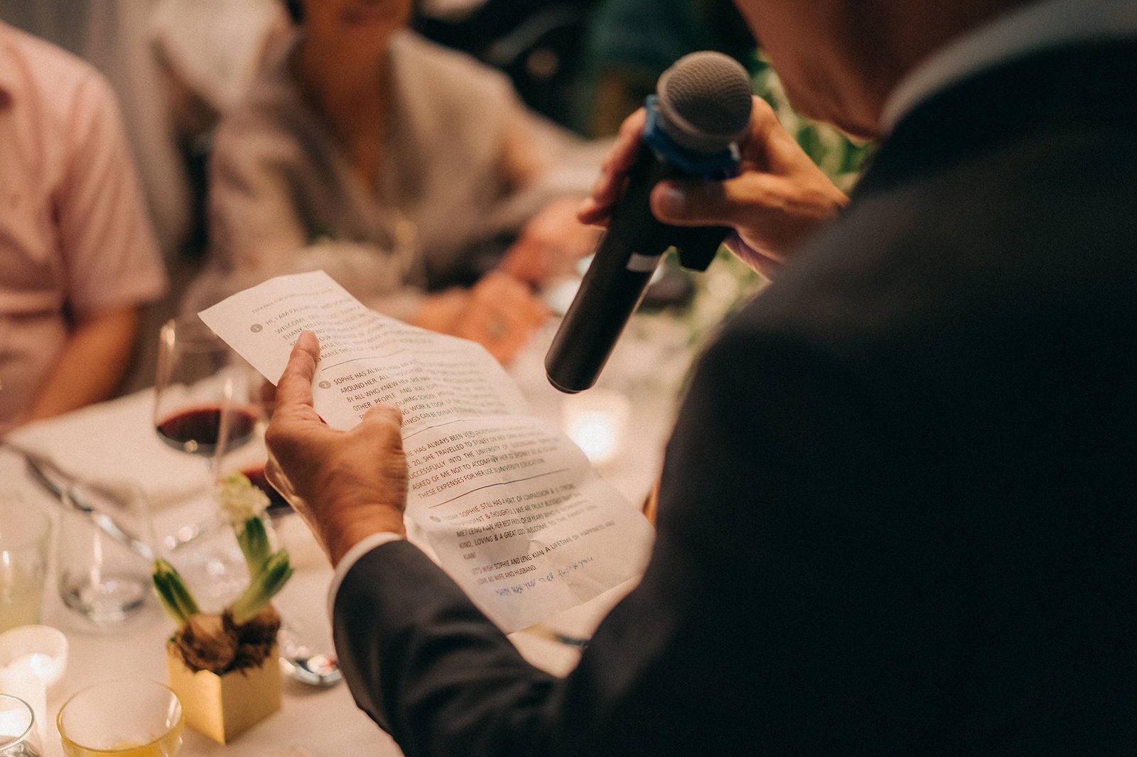 A man in a suit holds a microphone and a speech at a wedding reception at Monti at 1-Pavilion, Singapore, with guests blurred in the background.