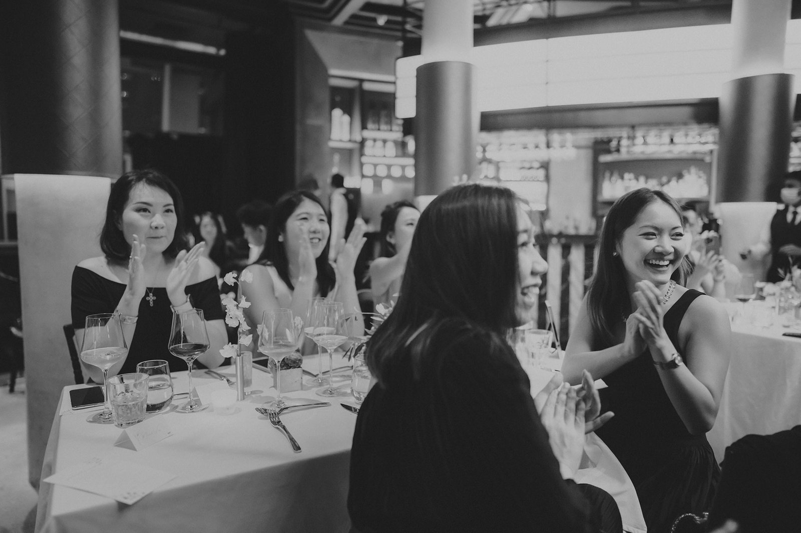 Guests clap and smile during a wedding reception at Monti at 1-Pavilion, Singapore, captured in black and white photography.