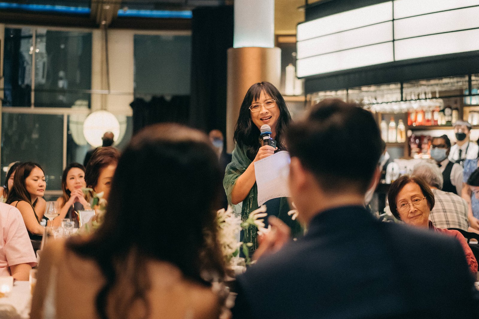 A woman with glasses and a green top speaks into a microphone at a wedding reception at Monti at 1-Pavilion, Singapore. Guests are seated at tables in the background, with soft lighting creating a warm atmosphere.