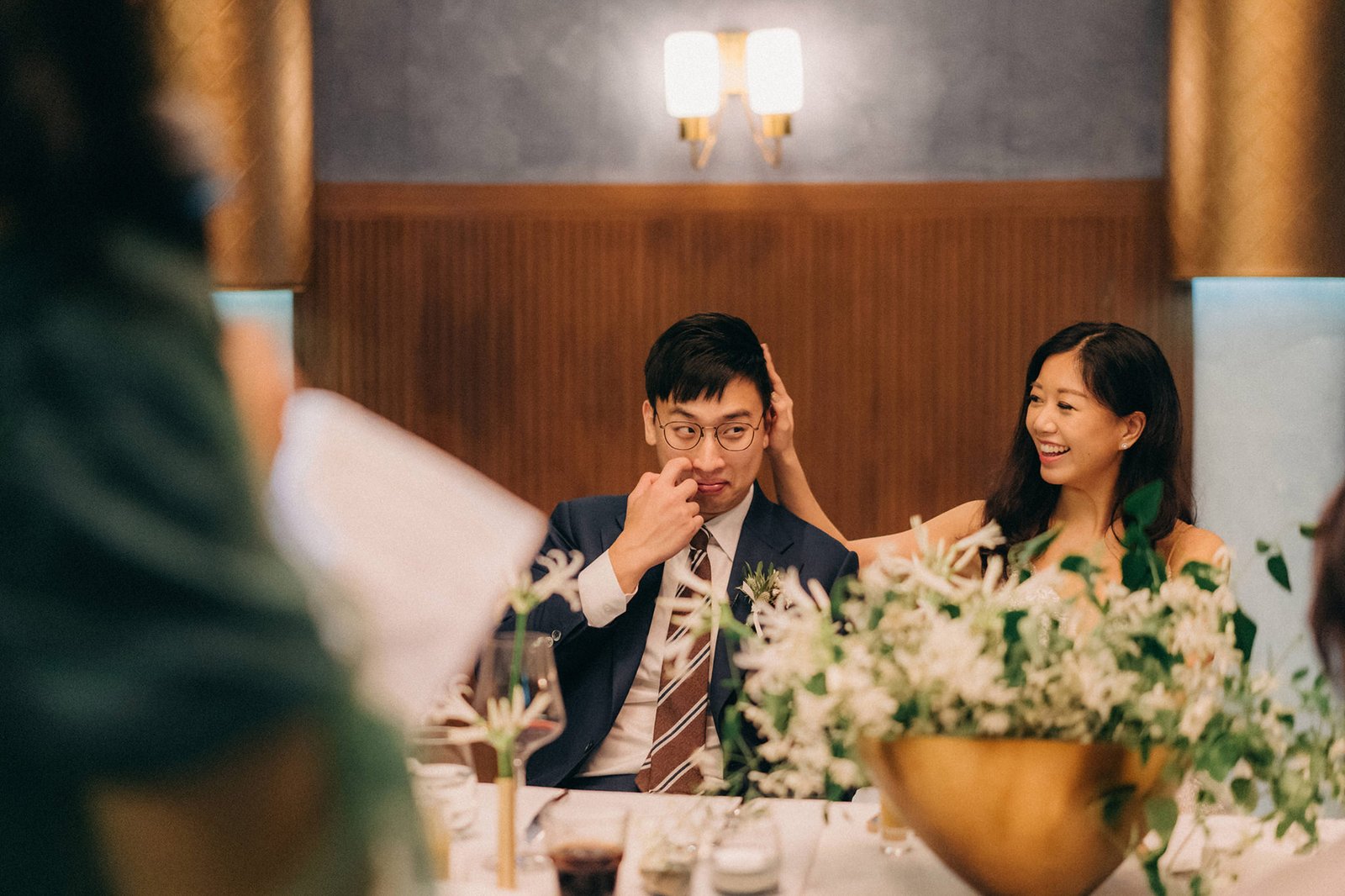 A laughing woman in a gold dress touches the head of a man in glasses and a suit at a wedding reception at Monti, Singapore. A floral centerpiece is in the foreground.