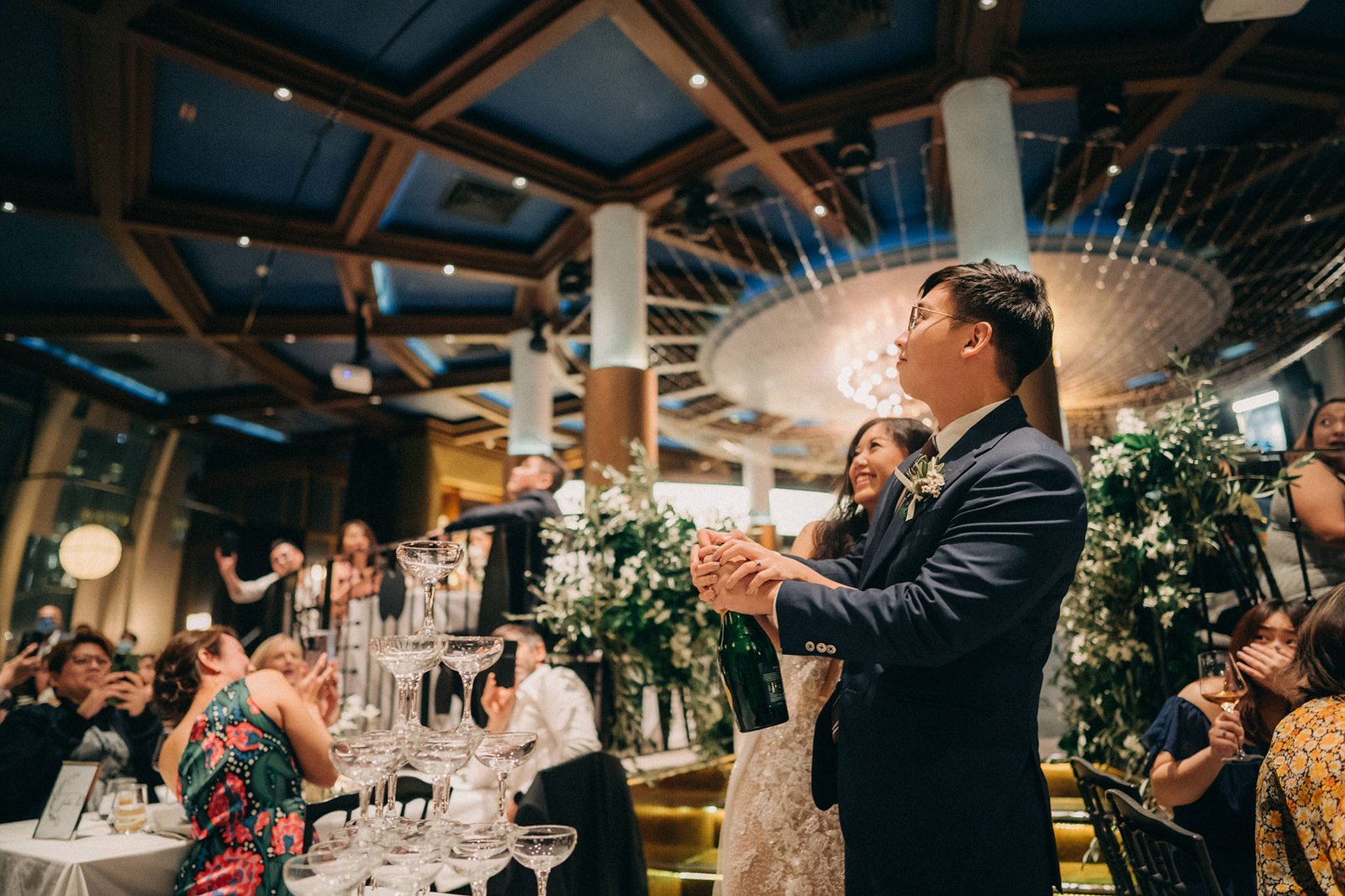 A groom in a navy suit opens a champagne bottle at a wedding reception at Monti, Singapore, with a champagne tower and guests in the background.