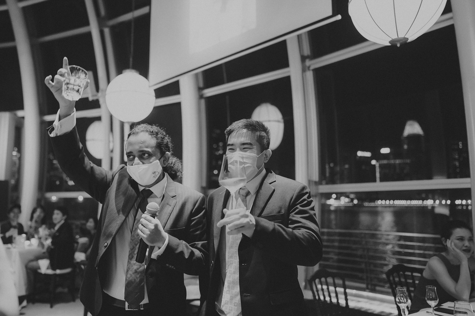 Two men in suits and face masks raise glasses at a wedding reception at Monti at 1-Pavilion, Singapore, with city lights visible through the windows.