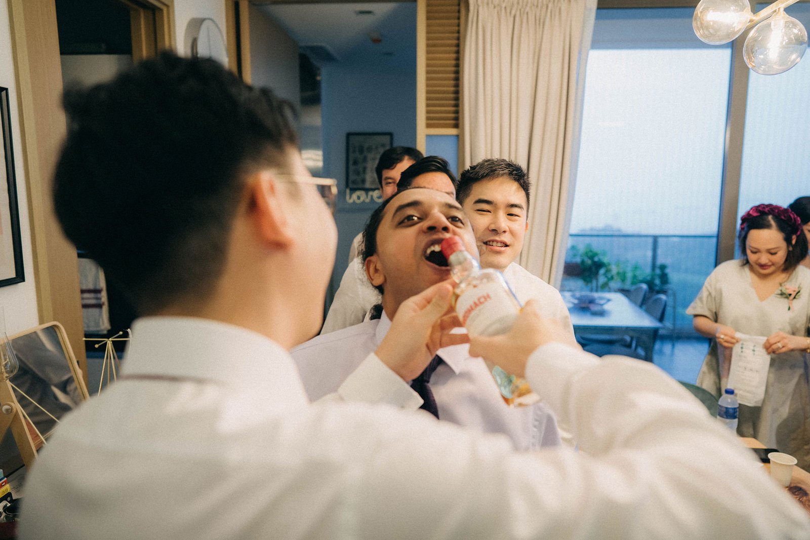 Grooming moment at a Monti wedding in Singapore: a groom is playfully offered a drink from a bottle by a friend during pre-wedding celebrations, with guests in the background.