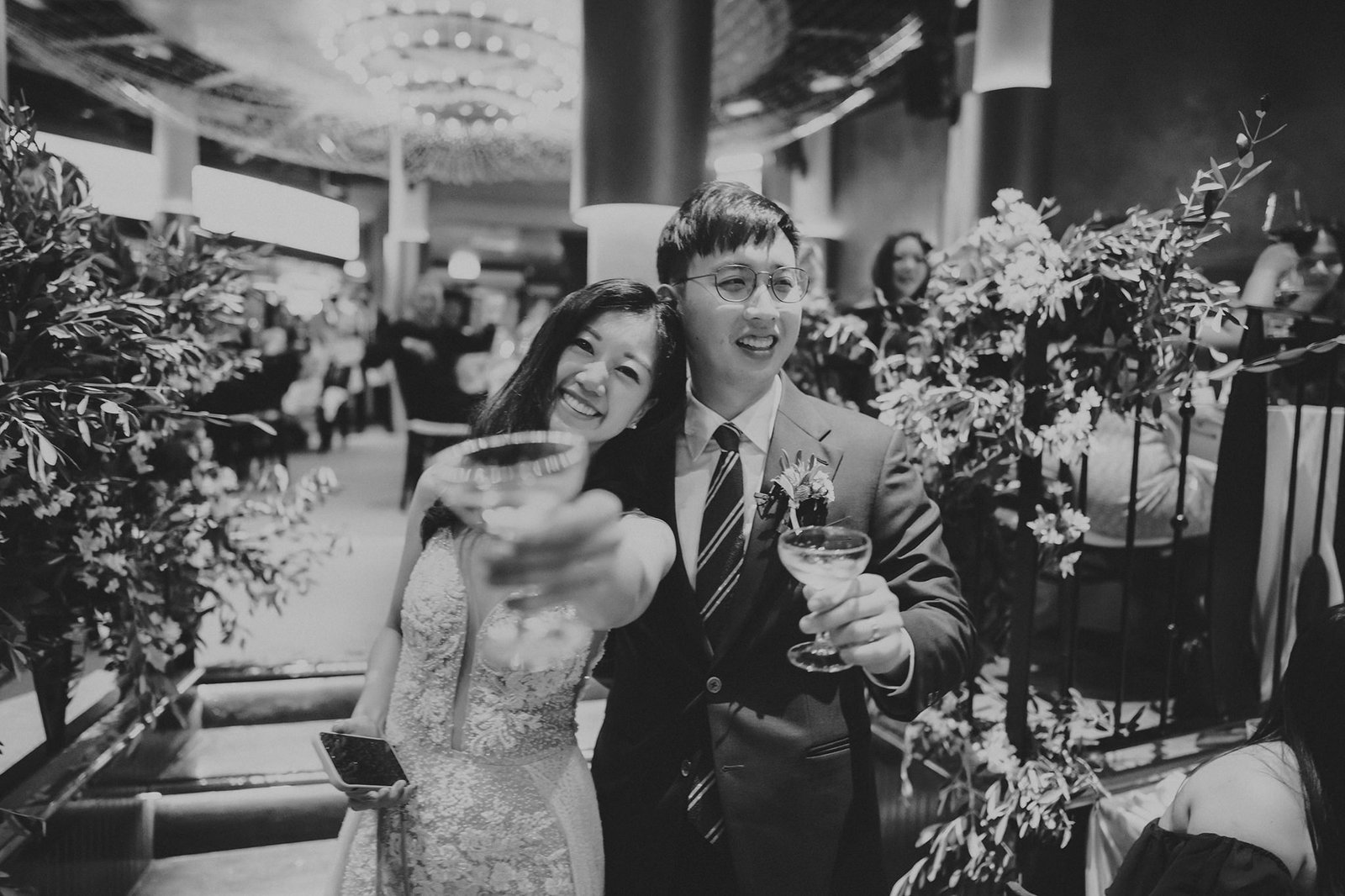 A smiling couple, LK and Sophie, toast with champagne glasses at their Monti at 1-Pavilion wedding in Singapore, captured in black and white photography.