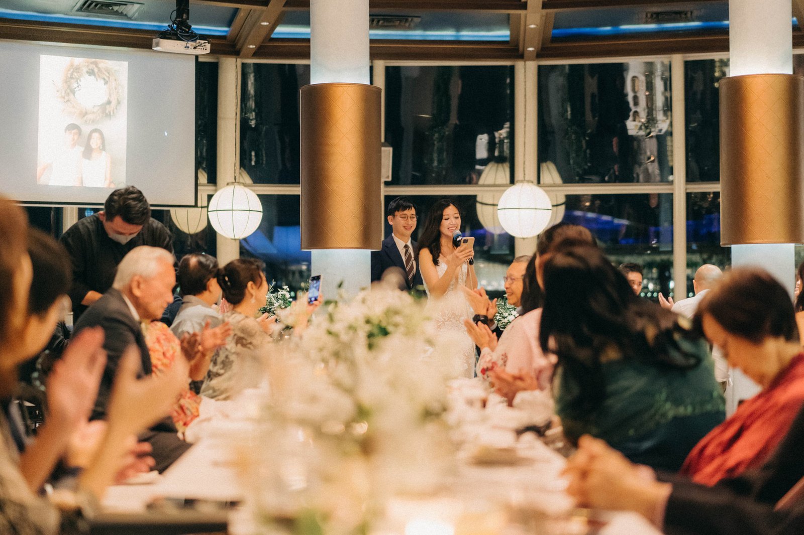 Bride and groom at their Monti at 1-Pavilion wedding reception in Singapore, with guests seated at a long table, illuminated by warm lighting and a projection screen.