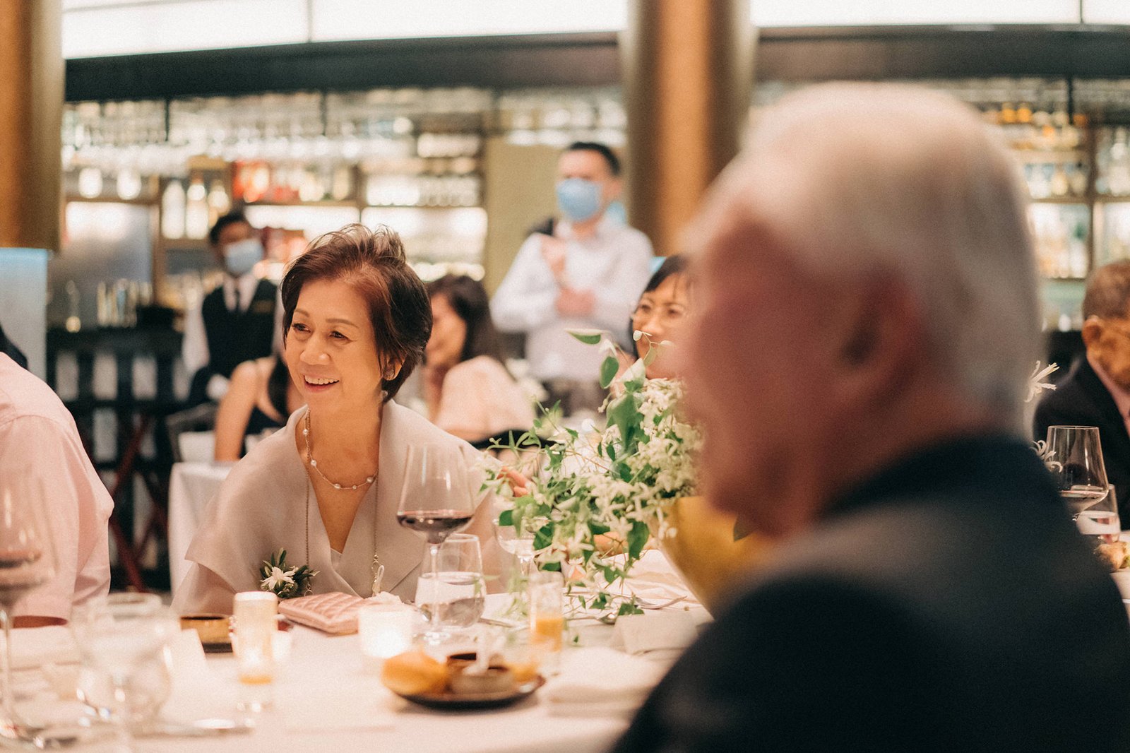 A smiling Asian woman in a light grey dress sits at a wedding reception table at Monti at 1-Pavilion, Singapore, with a bouquet of white flowers and a glass of red wine. Wedding photography captures the elegant atmosphere.