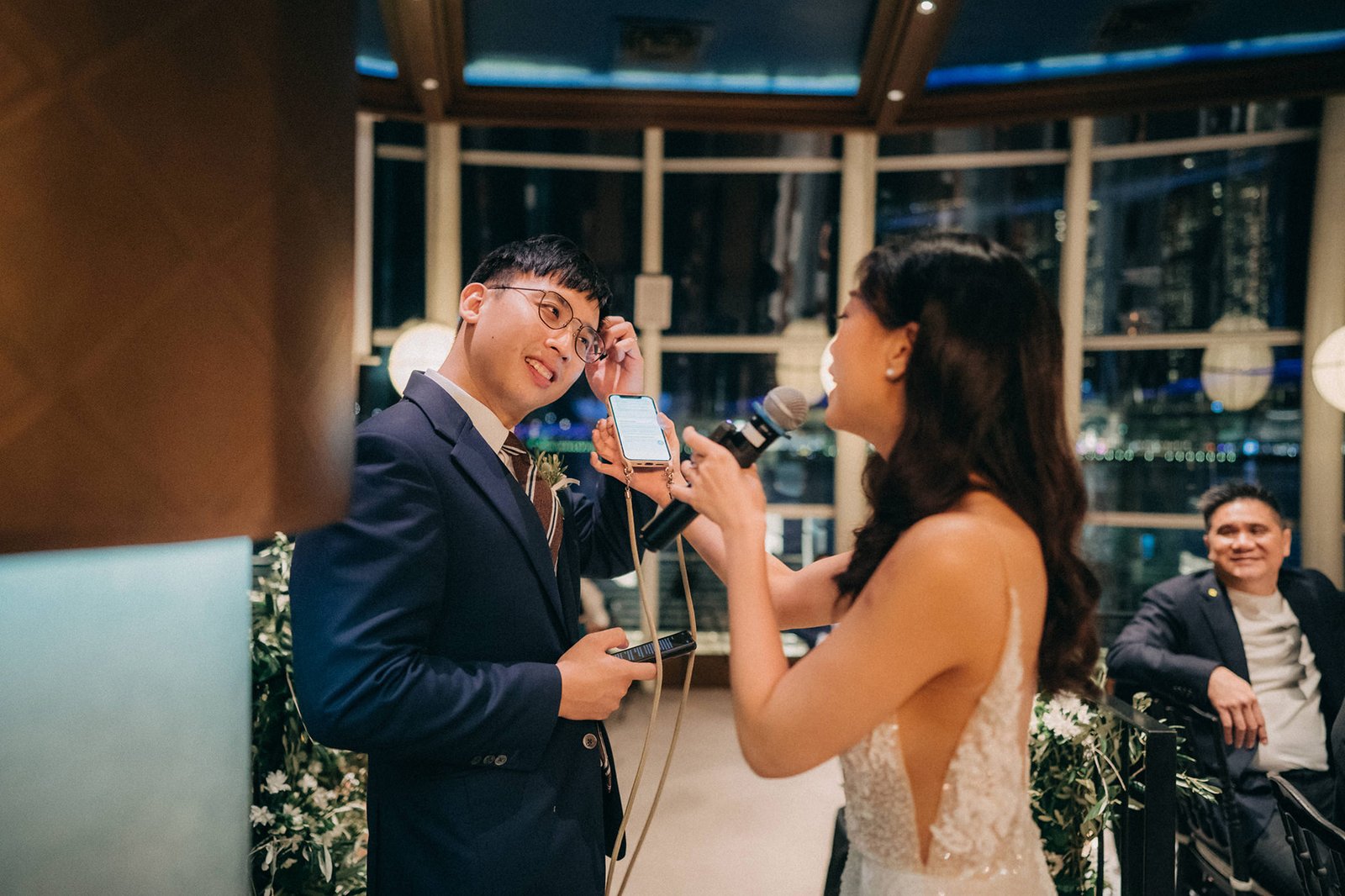 A groom in a navy suit holds up a phone to a bride in a white gown during a wedding reception at Monti at 1-Pavilion, Singapore. A man in a suit watches from the background.