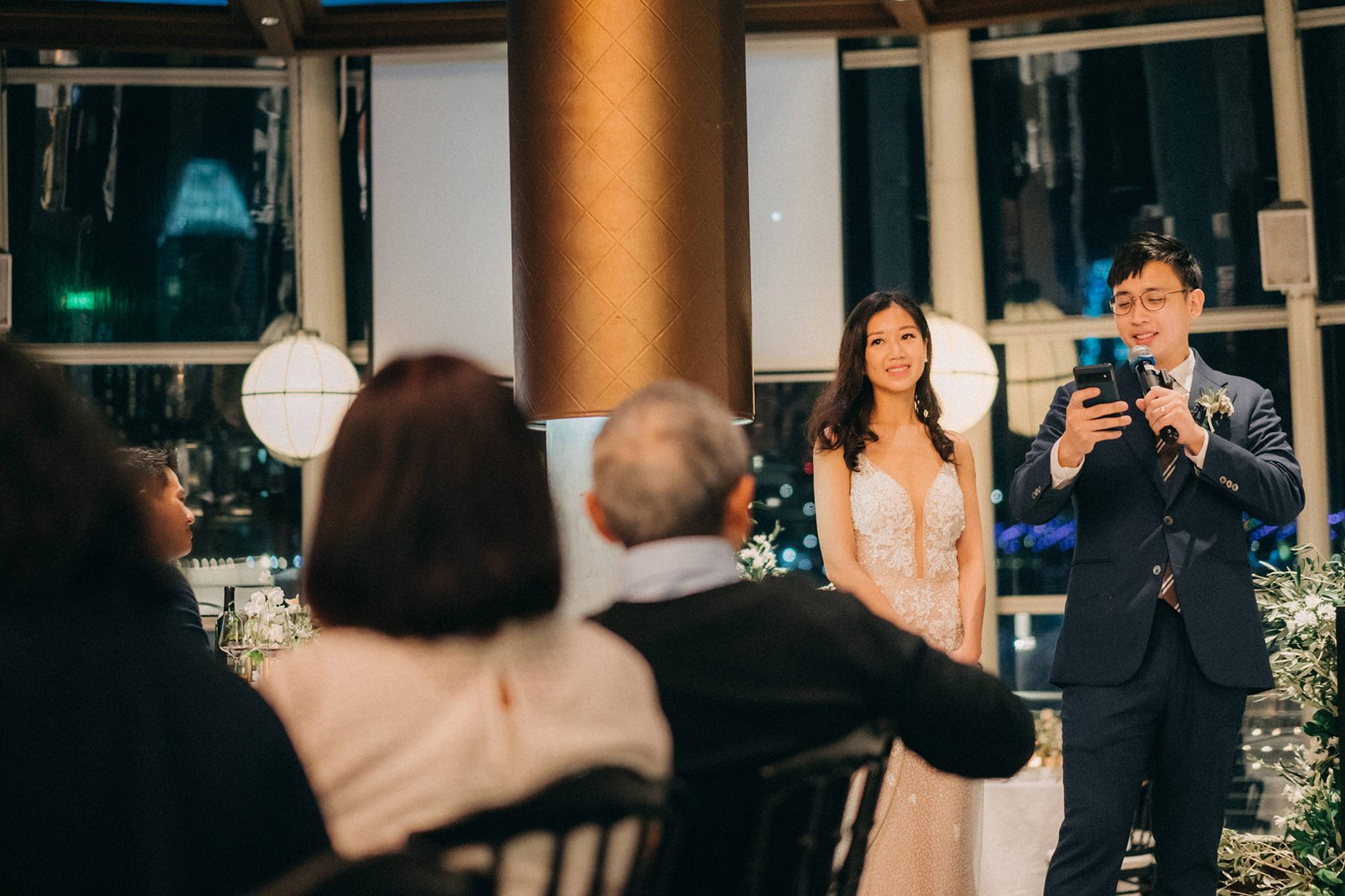 A bride in a white lace gown and groom in a navy suit speak at a wedding reception at Monti at 1-Pavilion, Singapore, with guests seated in the foreground.