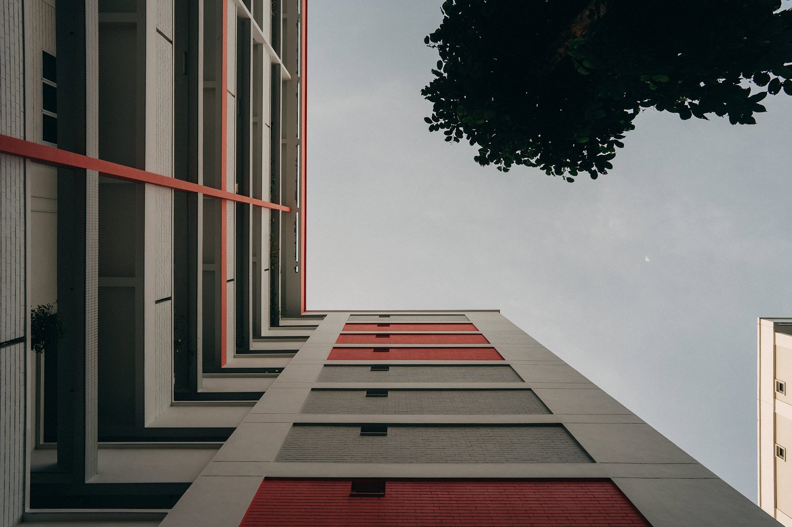 Low-angle view of modern apartment buildings with red and grey facades, seen from below against a pale sky with a tree branch overhead.