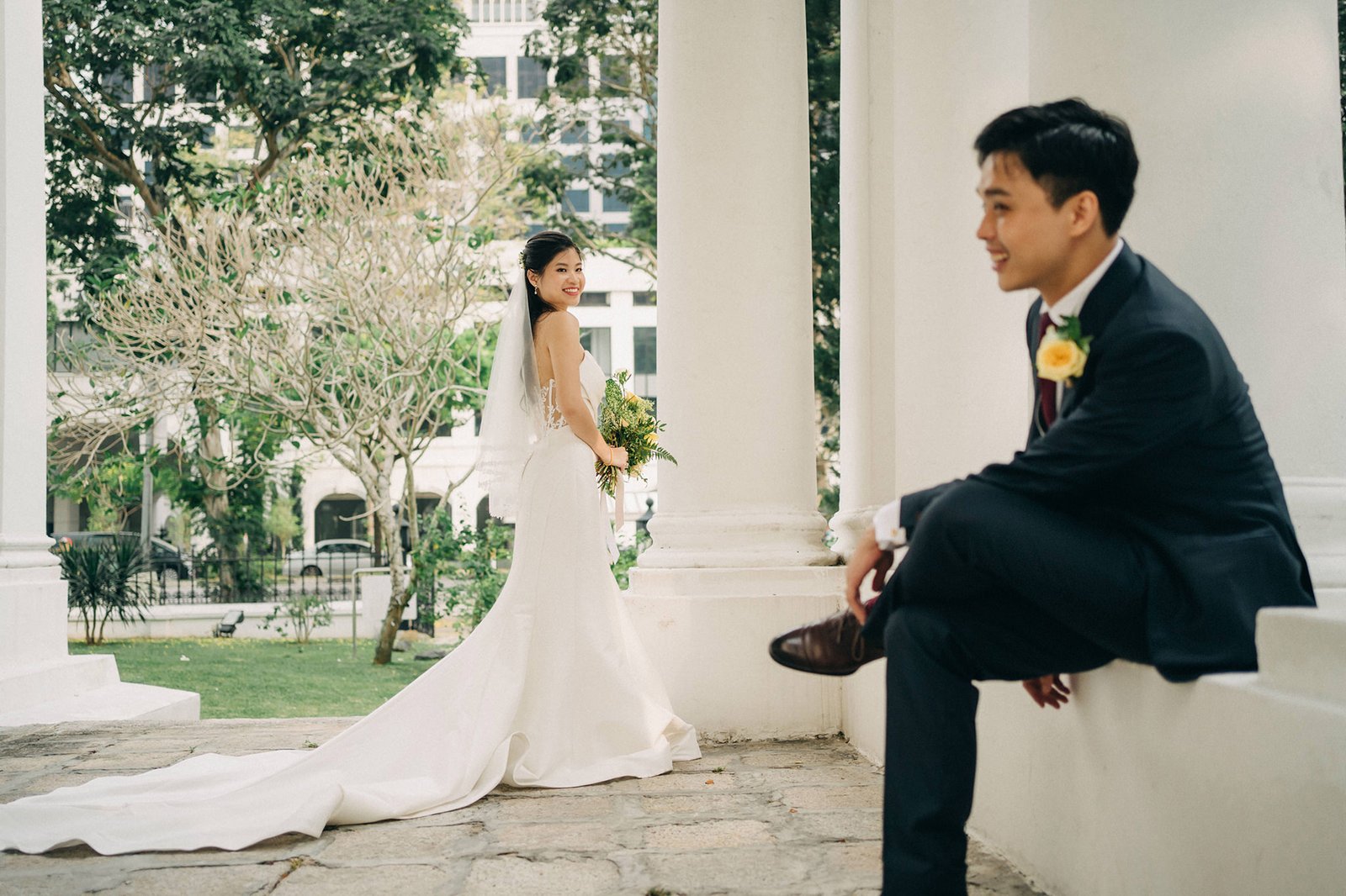 A smiling bride in a white wedding dress with a long train looks back at the groom, who is seated and looking at her, in a garden setting with white columns.